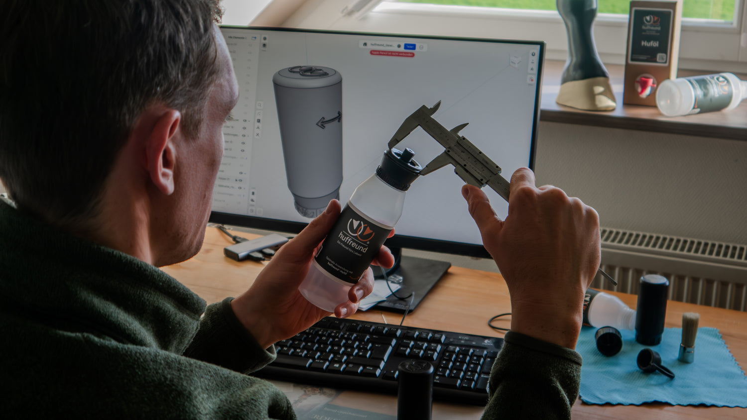 Ein Ingenieur sitzt mit einem Messchieber und der huffreund-Pinselflasche zur Auftragung von Huföl auf Pferdehufe vor einem Computer und gleicht Maße ab.