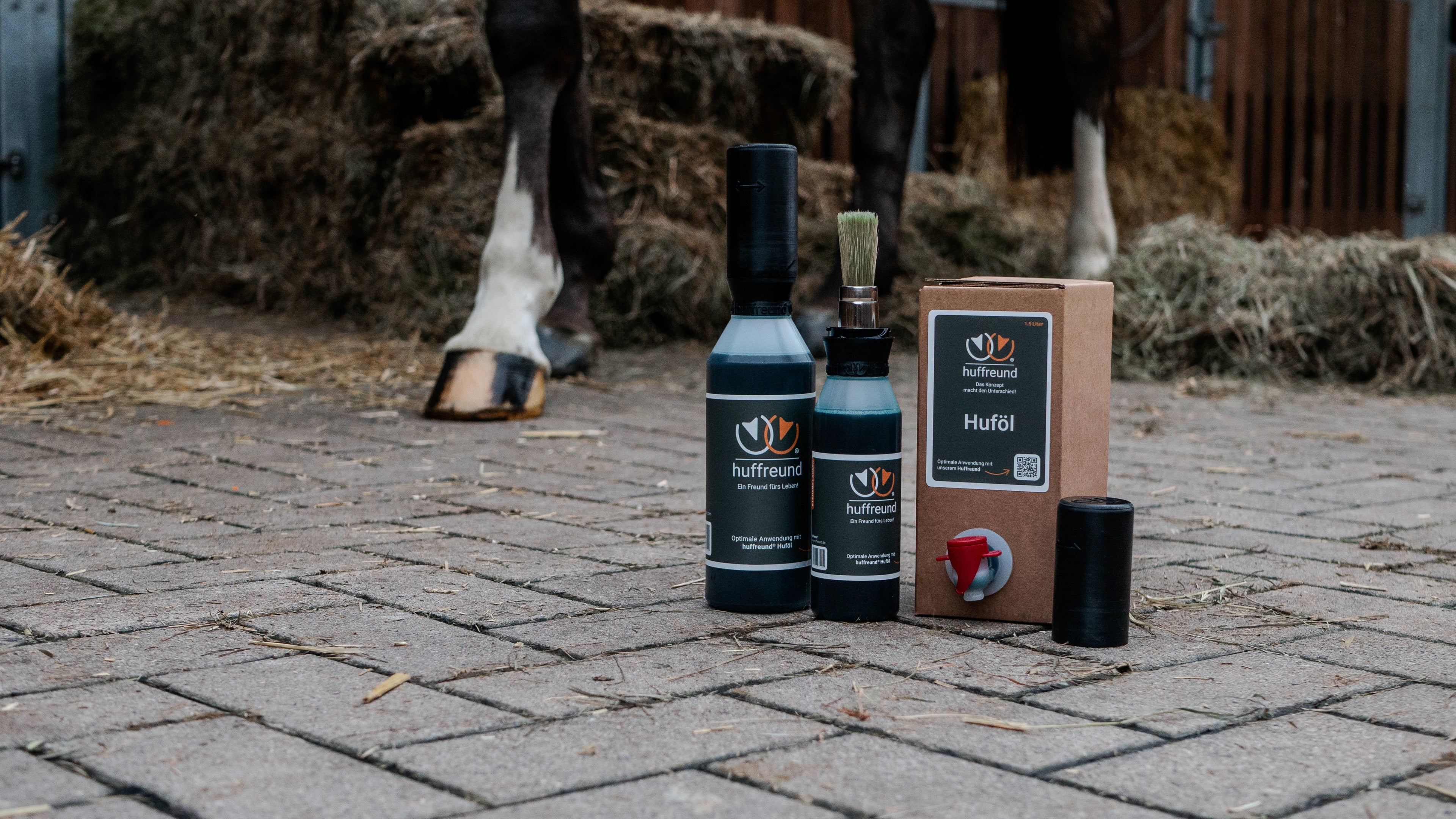 huffreund-Pinselflasche Pro und Compact sowie Huföl Bag-In-Box 1,5L stehen vor einem Pferdehuf im Hintergrund. 