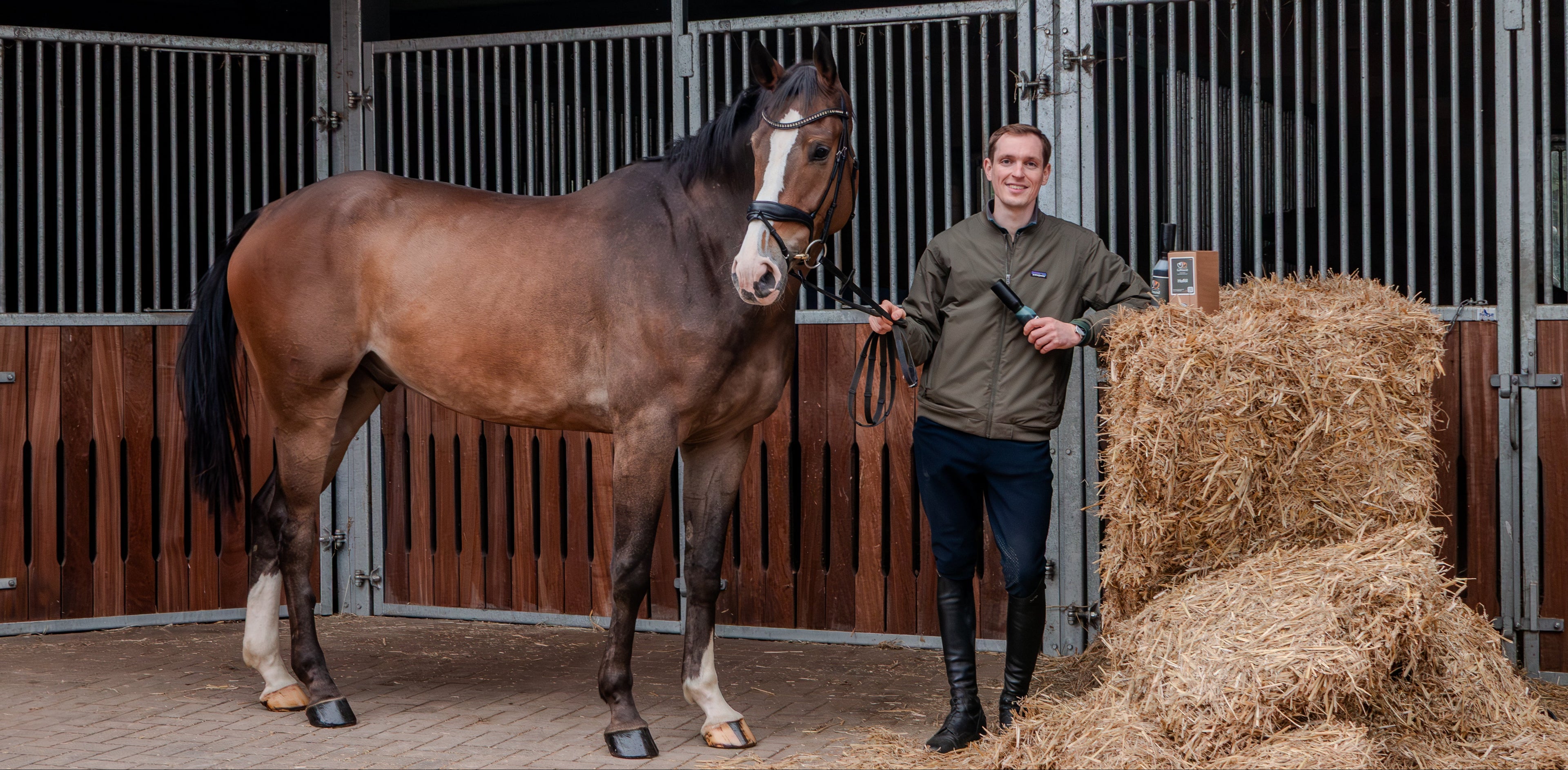 Der Unternehmensgründer steht mit seinem Pferd und den angebotenen Huföl Produkten vor Stalleinrichtungen im Hintergrund neben einem Strohbund.