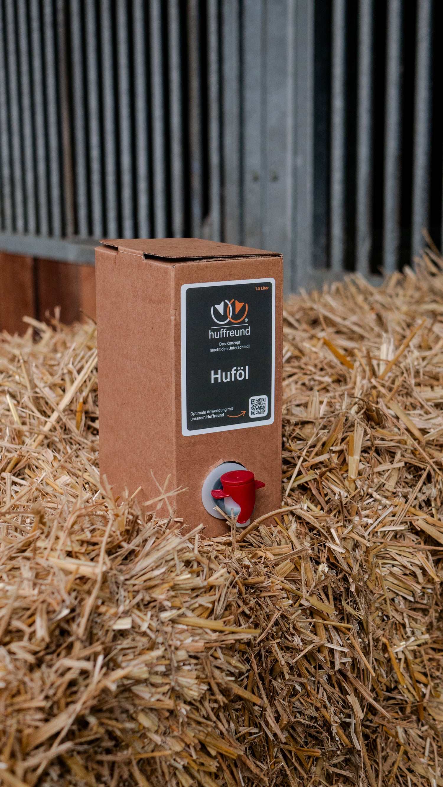 huffreund-Huföl für Pferde in Bag-In-Box 1,5L auf Stroh vor Stallungen im Hintergrund.
